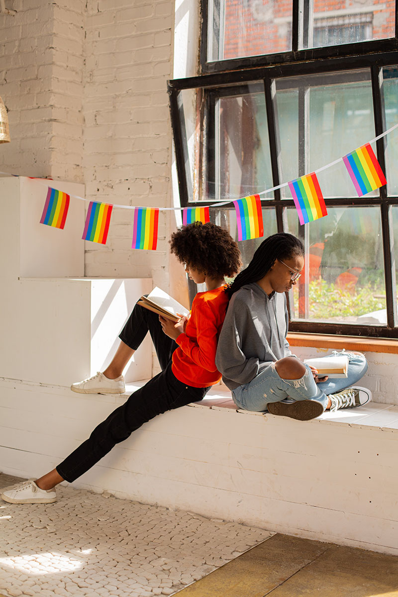 Two women reading under a rainbow banner by the window